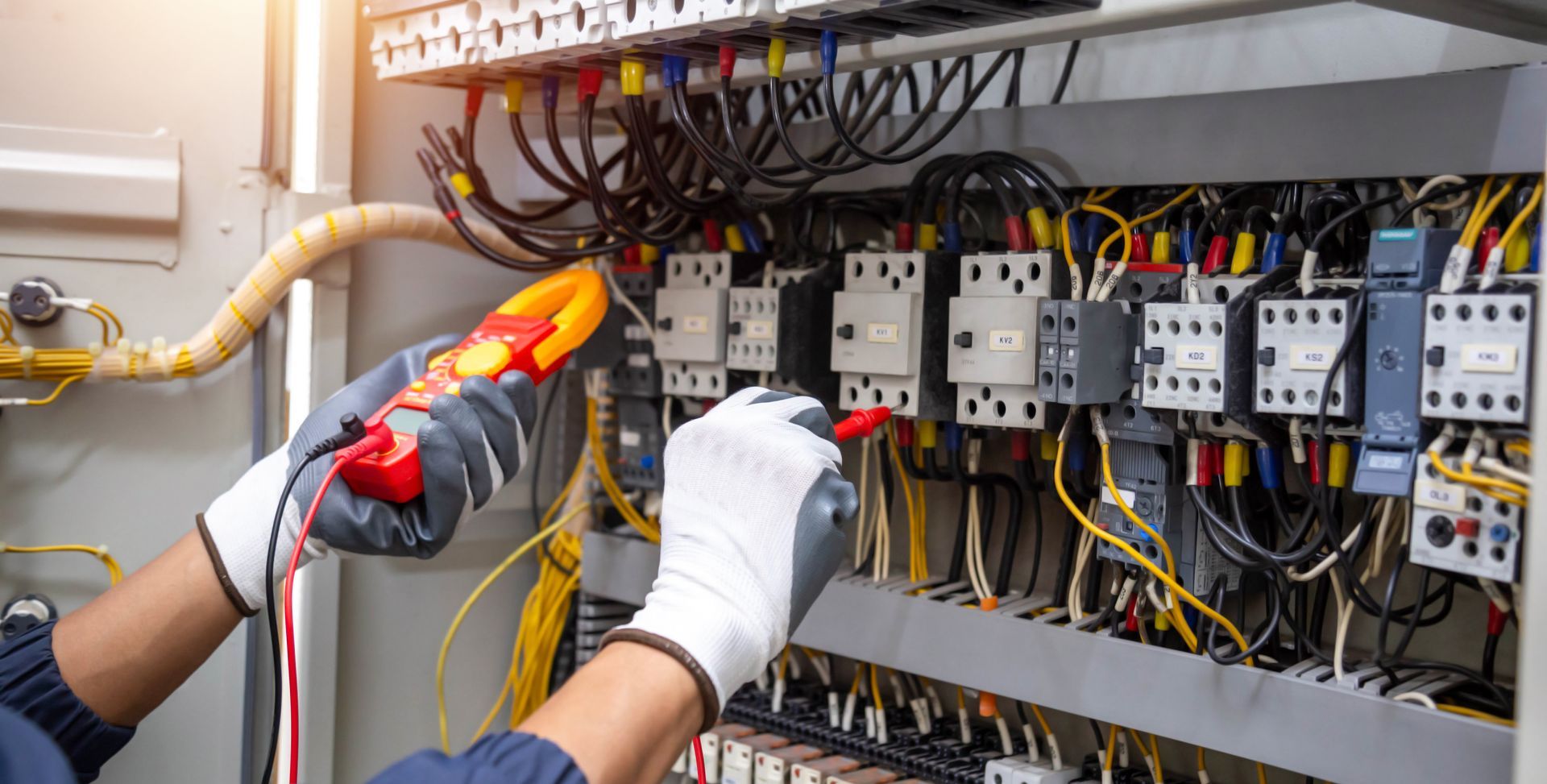 Un technicien portant des gants utilise un testeur de tension à l'intérieur d'un tableau électrique contenant des câbles et des disjoncteurs.