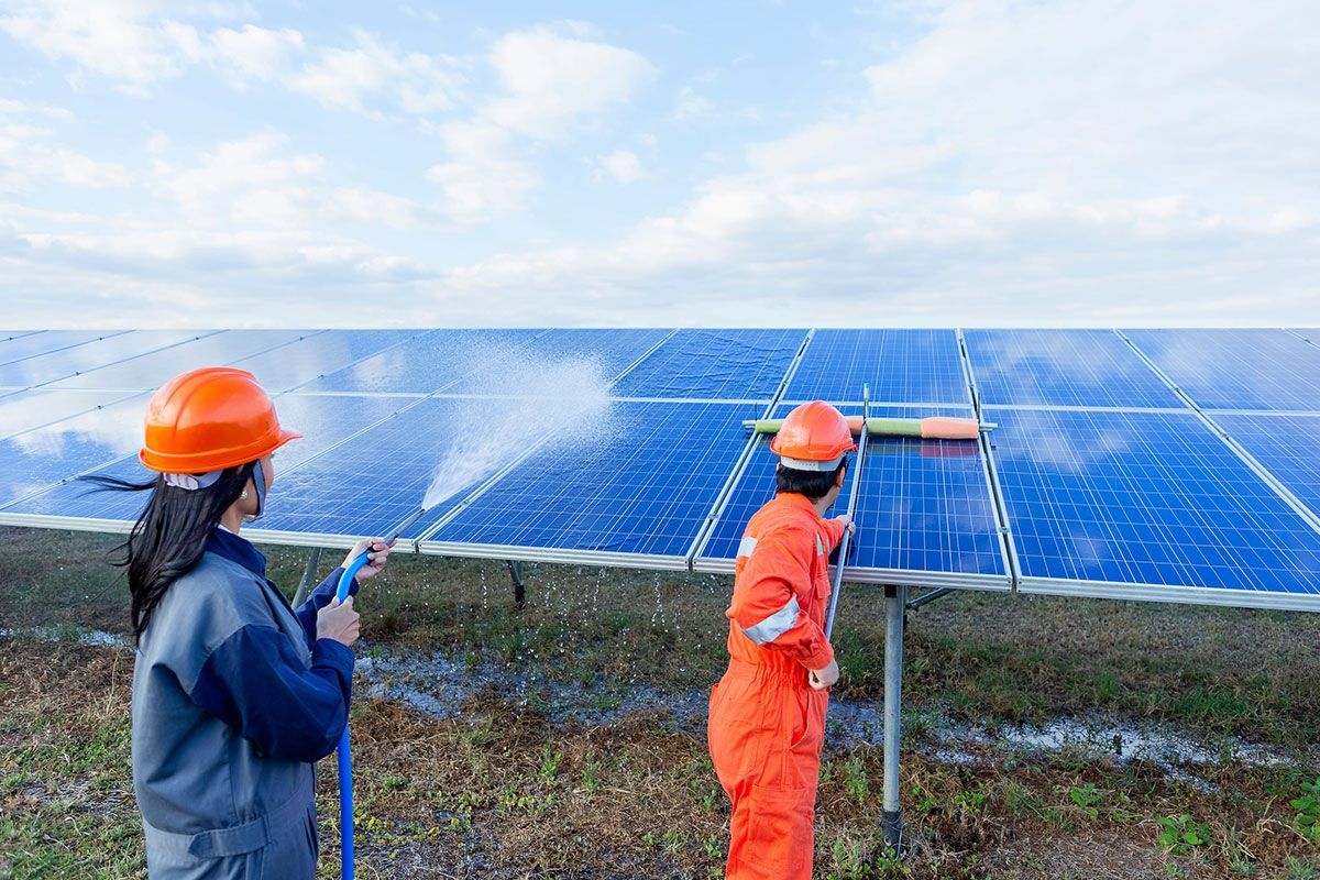 Dos trabajadores con equipo de seguridad limpian paneles solares con un pulverizador de agua y una escobilla de goma en un campo al aire libre.