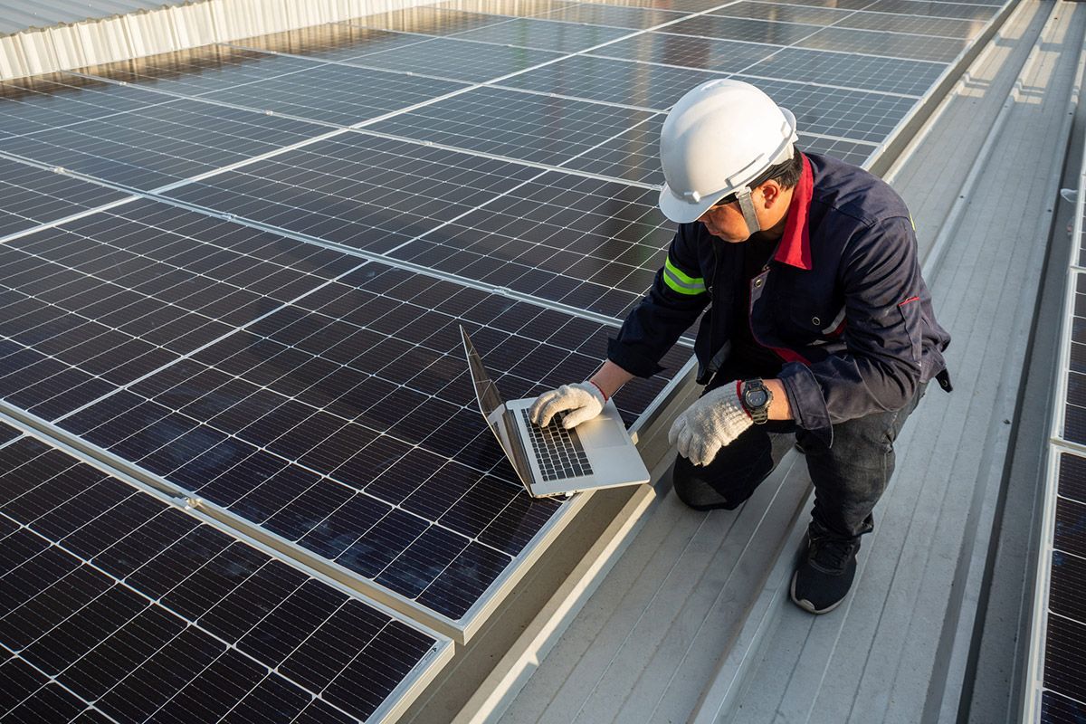 Un técnico, con casco blanco y ropa de trabajo, está arrodillado en un tejado revisando los paneles solares con un ordenador portátil.