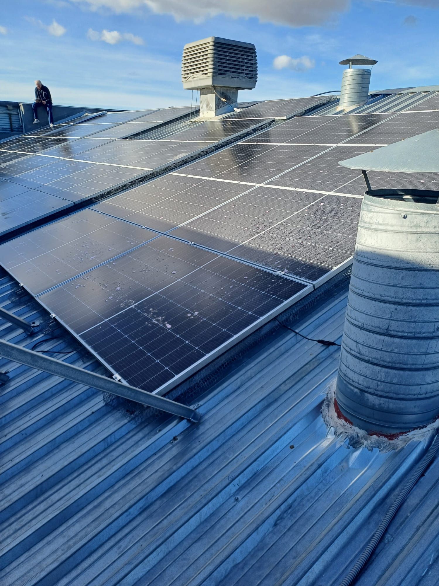 Paneles solares instalados en un tejado metálico con chimenea, ventilación y enfriador evaporativo en un día soleado y parcialmente nublado.