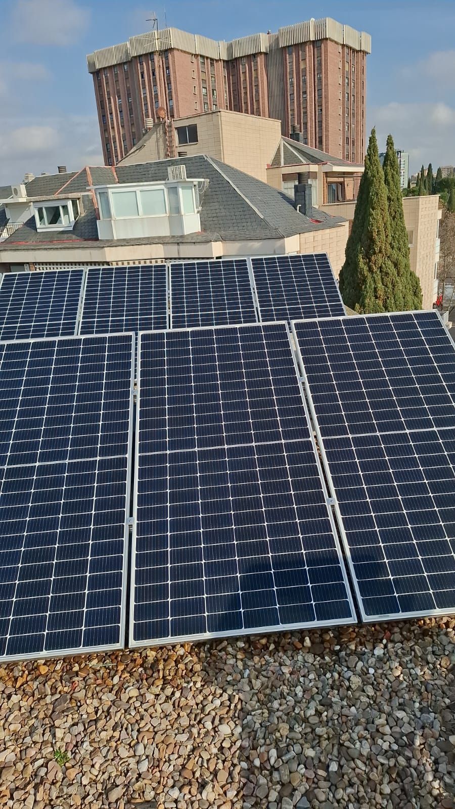 Un conjunto de paneles solares instalados en una azotea, dispuestos en forma de cuadrícula, con un edificio y árboles al fondo bajo un cielo azul nublado.