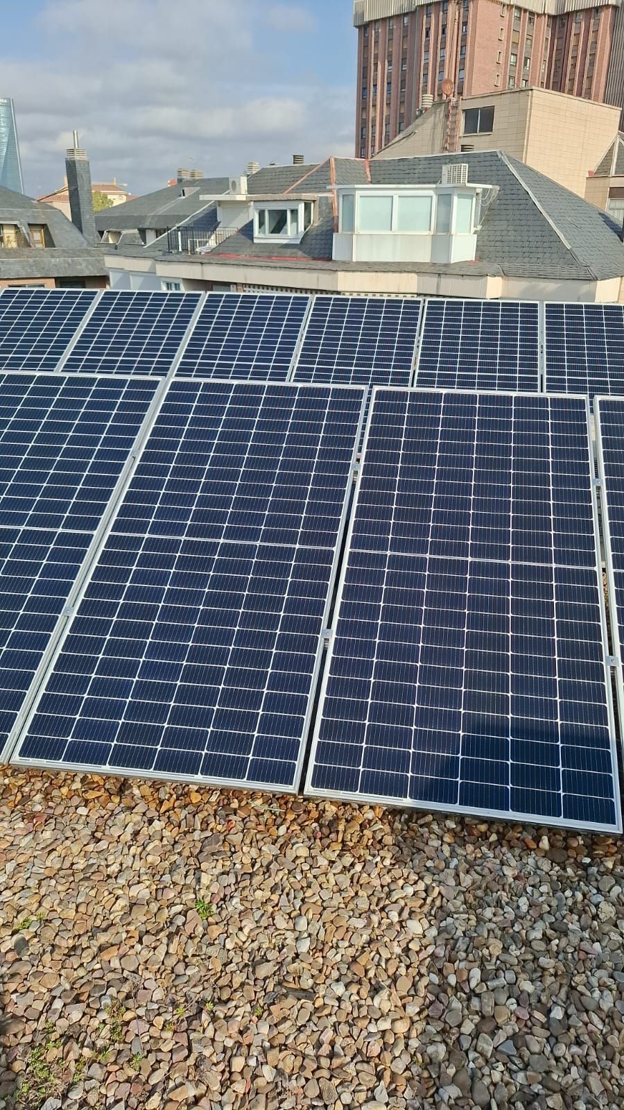 Paneles solares instalados en un tejado plano cubierto de grava, con casas visibles al fondo bajo un cielo azul.