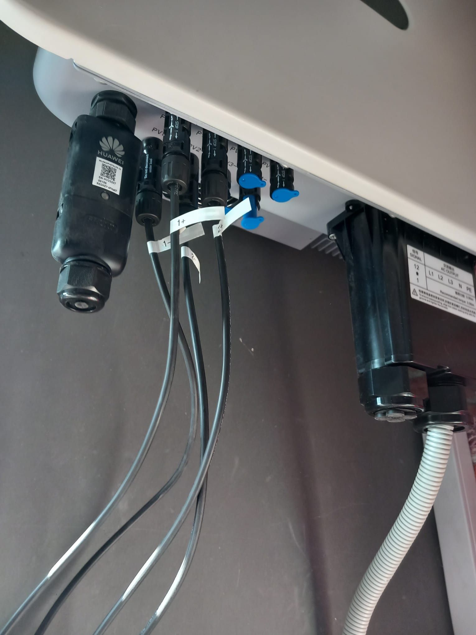 Vista inferior de un inversor solar de montaje en pared con cables eléctricos negros conectados y puertos de entrada con tapas azules.