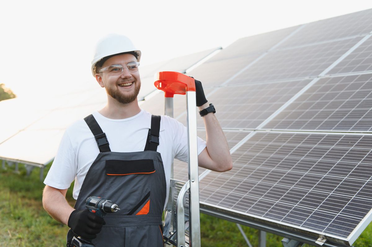 Un trabajador con equipo de protección y casco sostiene un taladro mientras está de pie junto a unos paneles solares en un campo al aire libre.