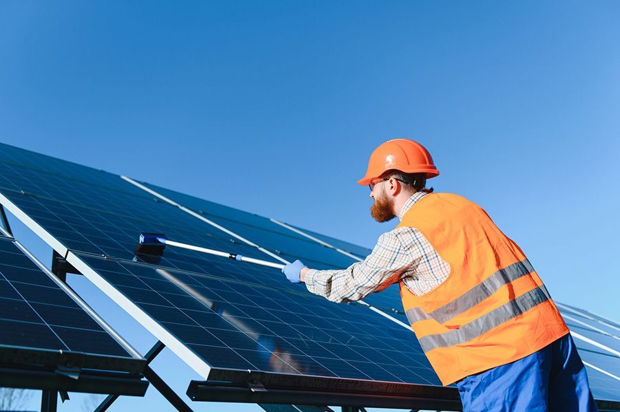 Un trabajador con chaleco de seguridad naranja y casco limpia paneles solares bajo un cielo azul despejado.