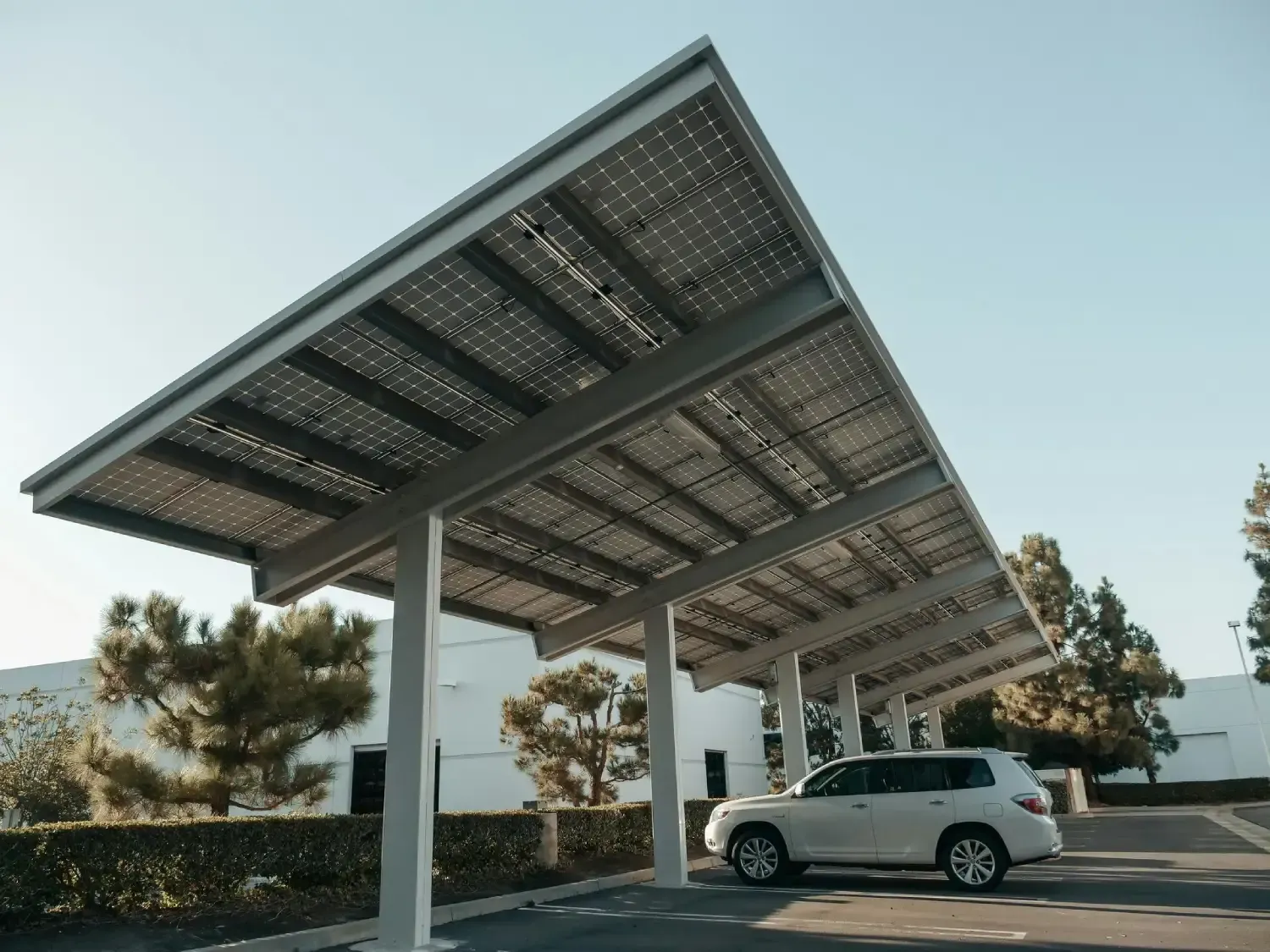 Cochera con paneles solares sobre una camioneta blanca estacionada. Vigas de acero, paneles y un soleado entorno exterior.