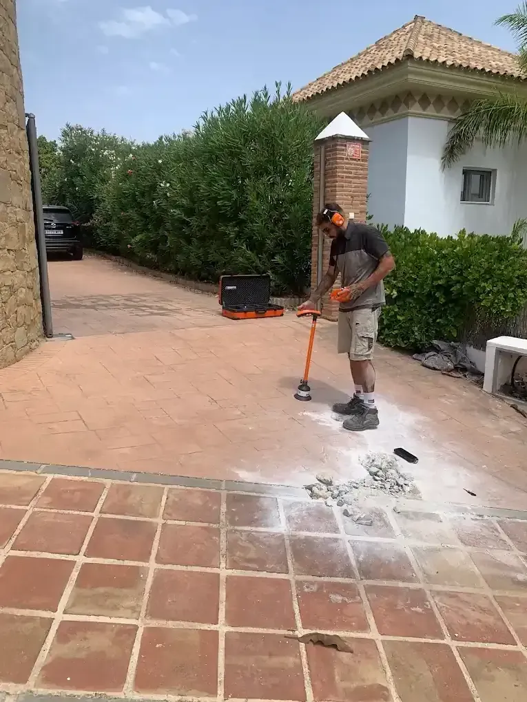 Un hombre usa una herramienta eléctrica en un patio de baldosas rojas al aire libre. Hay polvo y escombros.