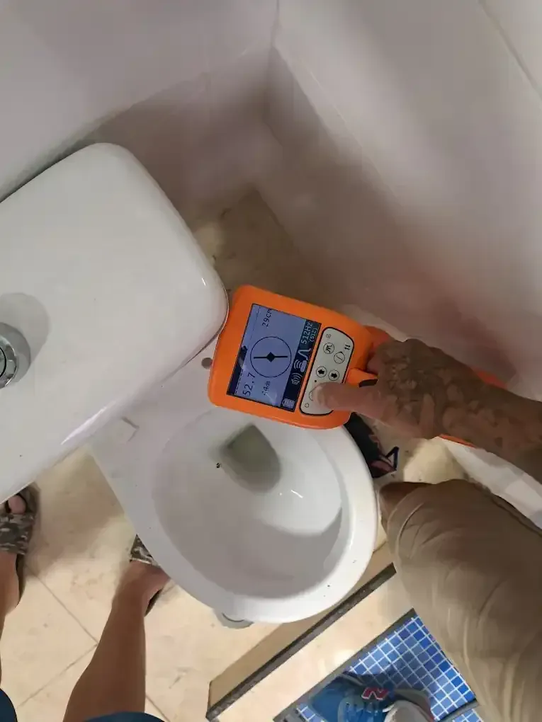 Persona que utiliza un dispositivo naranja para probar un inodoro blanco en un baño con azulejos de color beige.