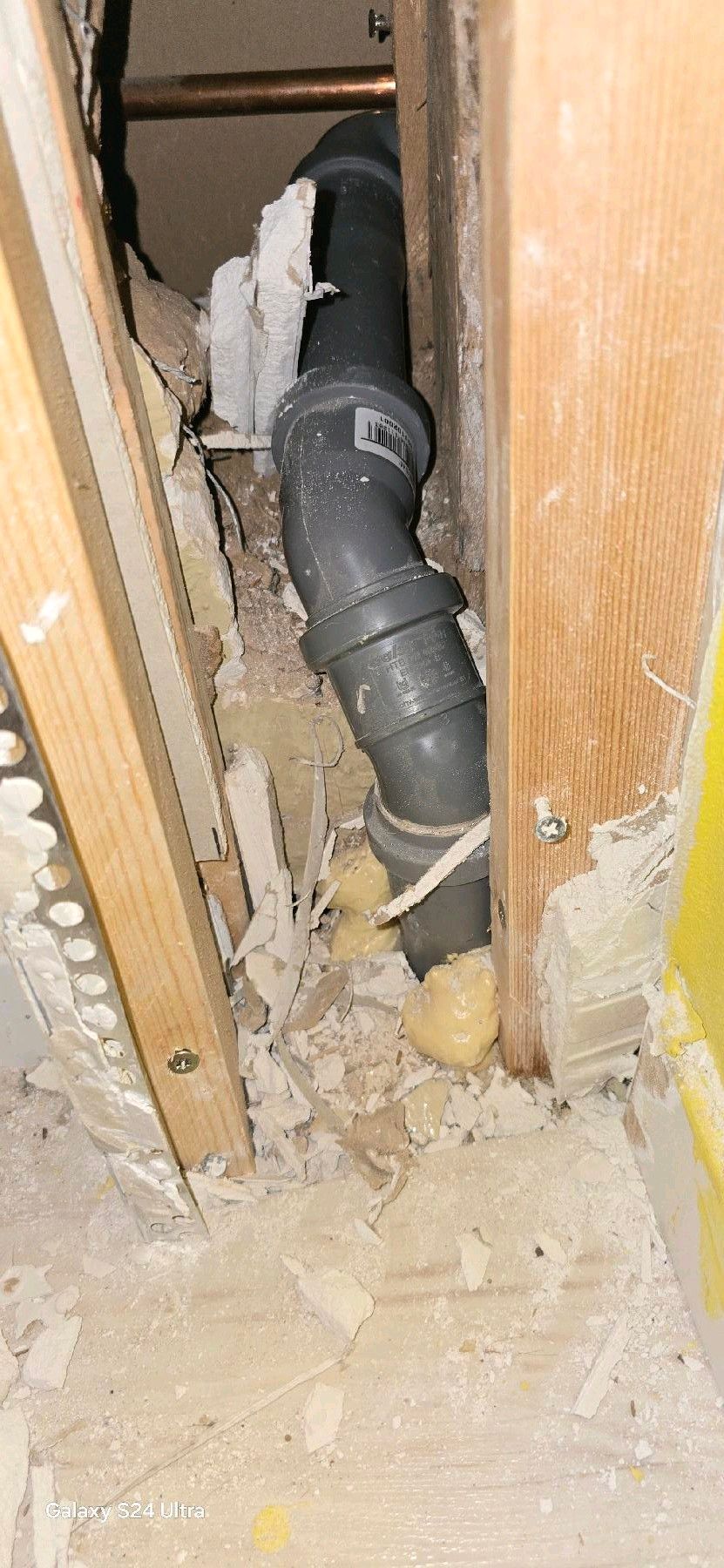 Ein dunkelgraues Rohr in einem Wandhohlraum, inmitten freiliegender Holzrahmen und Schutt.