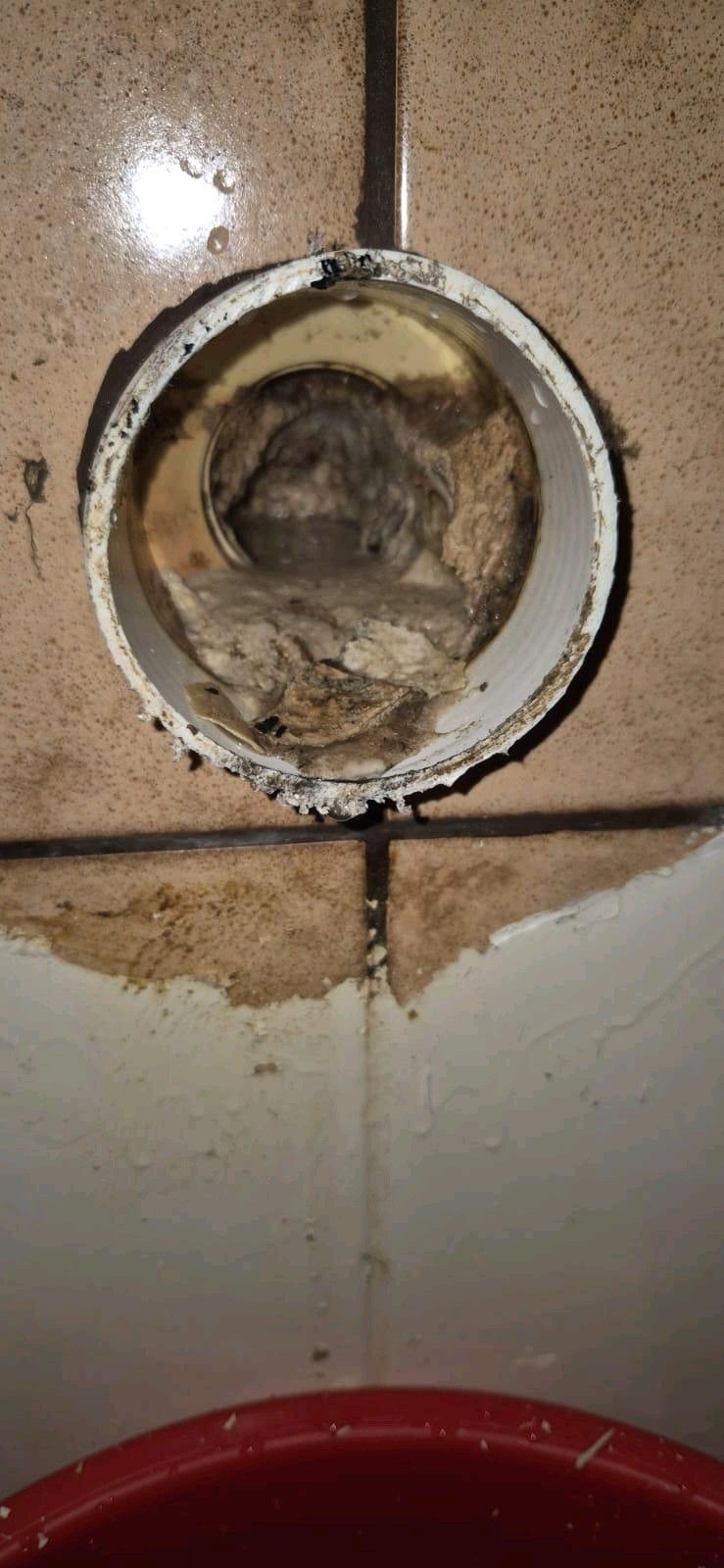 Ein PVC-Rohr in einer gefliesten Wand mit Schutt im Inneren und Flecken darunter. Ein roter Eimer ist teilweise sichtbar.