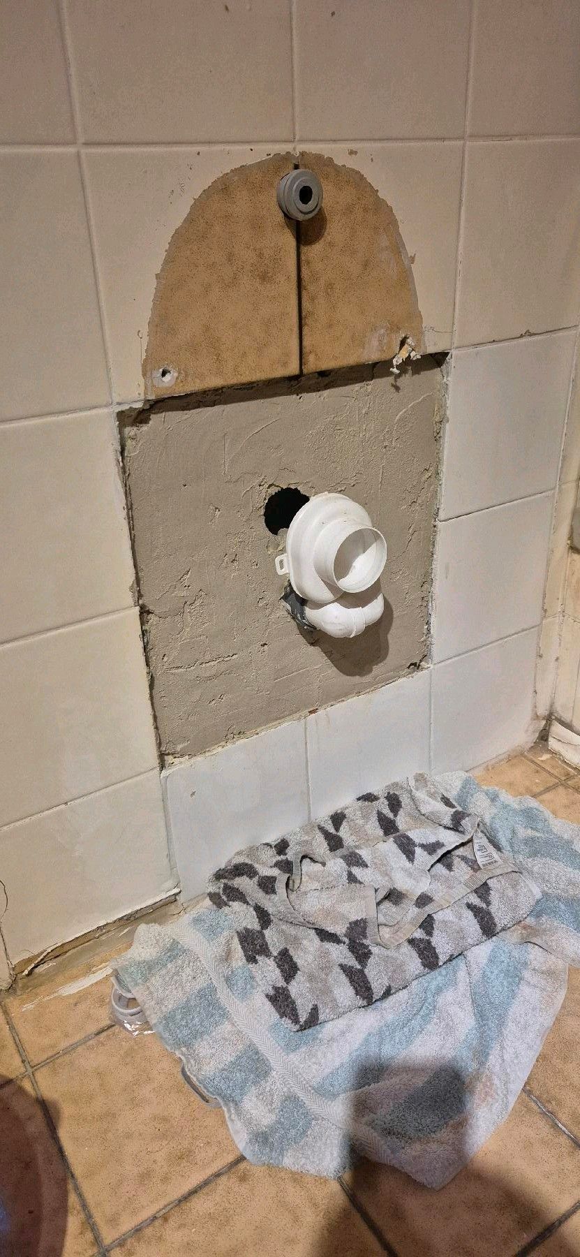 Badezimmerwand mit freiliegenden Rohrleitungen, ein Handtuch auf dem Boden.