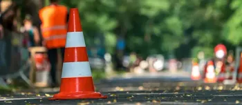 Orange-weißer Verkehrskegel auf einer Straße, unscharfer Hintergrund mit Menschen und Grün.