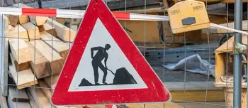 Bauwarnschild eines Arbeiters in einem roten Dreieck mit Holzbrettern und Drahtzaun im Hintergrund.