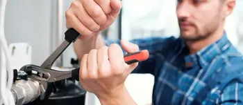 Ein Klempner bearbeitet ein Rohr mit einer Zange. Er trägt ein blau kariertes Hemd.