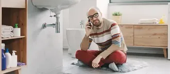 Mann im Badezimmer. Er sitzt mit einem Telefon auf dem Boden und sieht besorgt aus. Waschbecken und Leitungen sind sichtbar.