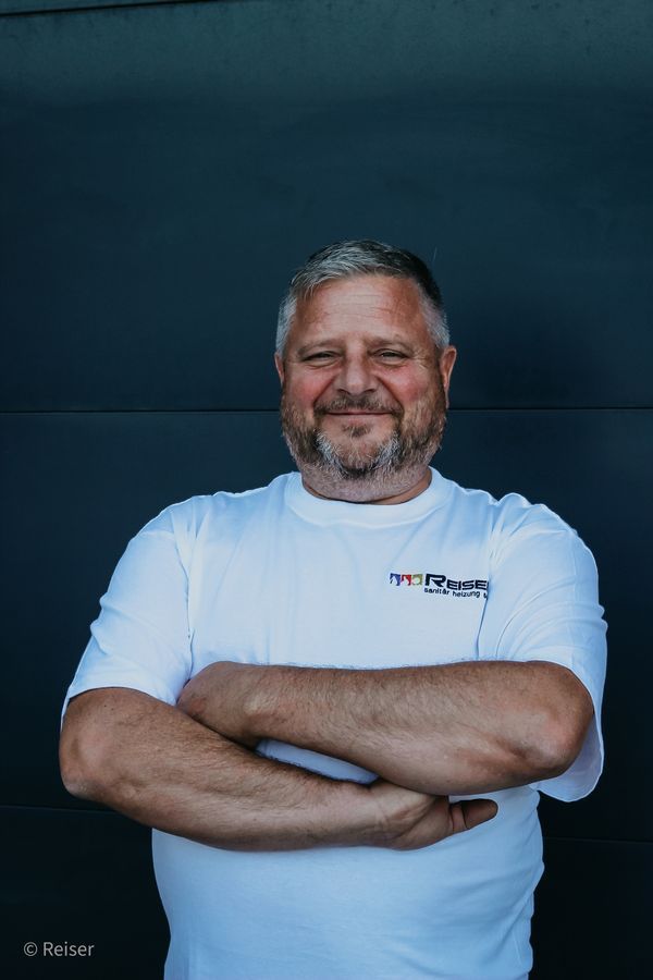 Mann in weißem T-Shirt mit Logo, die Arme verschränkt, lächelt vor einer dunkelgrauen Wand.
