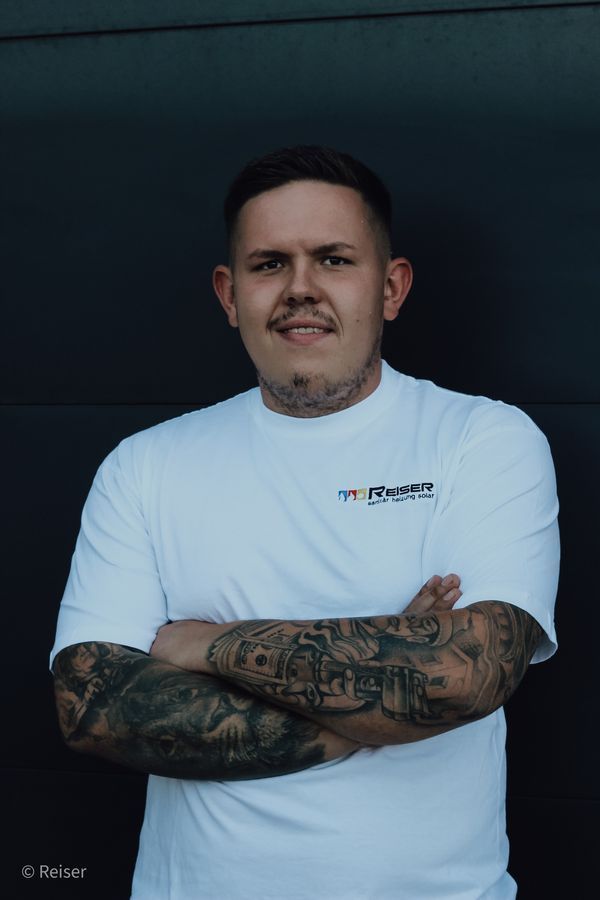 Mann mit Tätowierungen, weißes Hemd, verschränkte Arme, lächelnd. Dunkler Hintergrund.