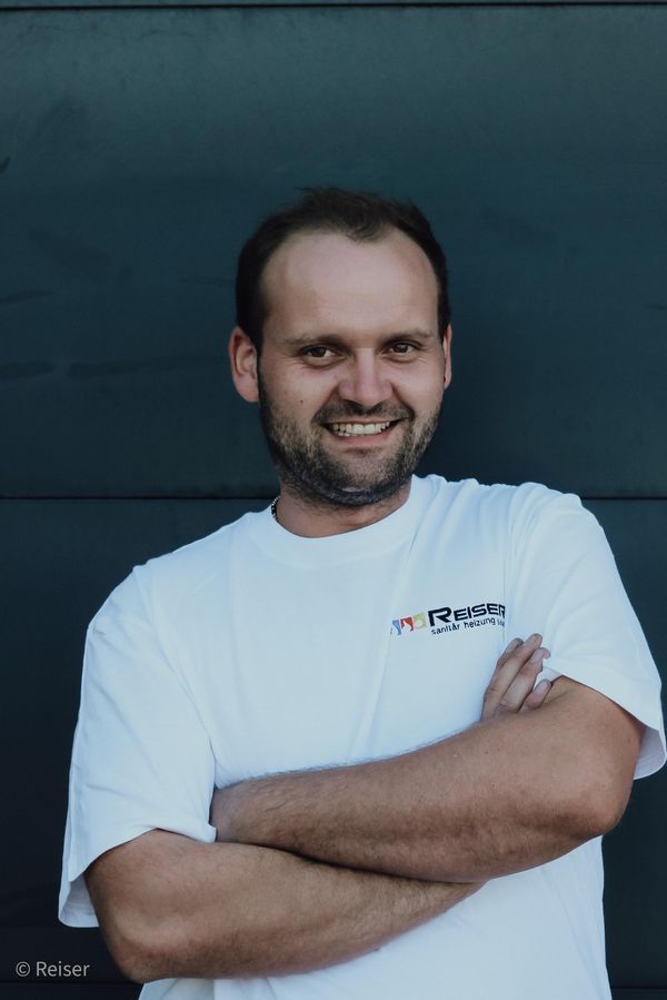 Mann in weißem T-Shirt mit Logo, lächelnd, die Arme verschränkt. Steht vor einer dunklen, massiven Wand.