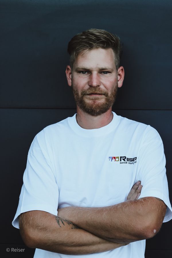 Ein Mann mit verschränkten Armen, der ein weißes T-Shirt trägt, steht vor einer dunklen Wand.