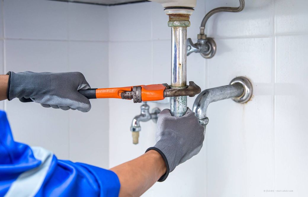 In Arbeitshandschuhen wird mit einem Schraubenschlüssel an den Wasserleitungen unter einem Waschbecken in einem weiß gefliesten Raum justiert.