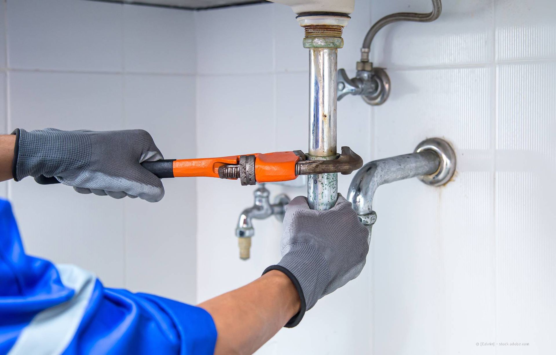 In Arbeitshandschuhen wird mit einem Schraubenschlüssel an den Wasserleitungen unter einem Waschbecken in einem weiß gefliesten Raum justiert.