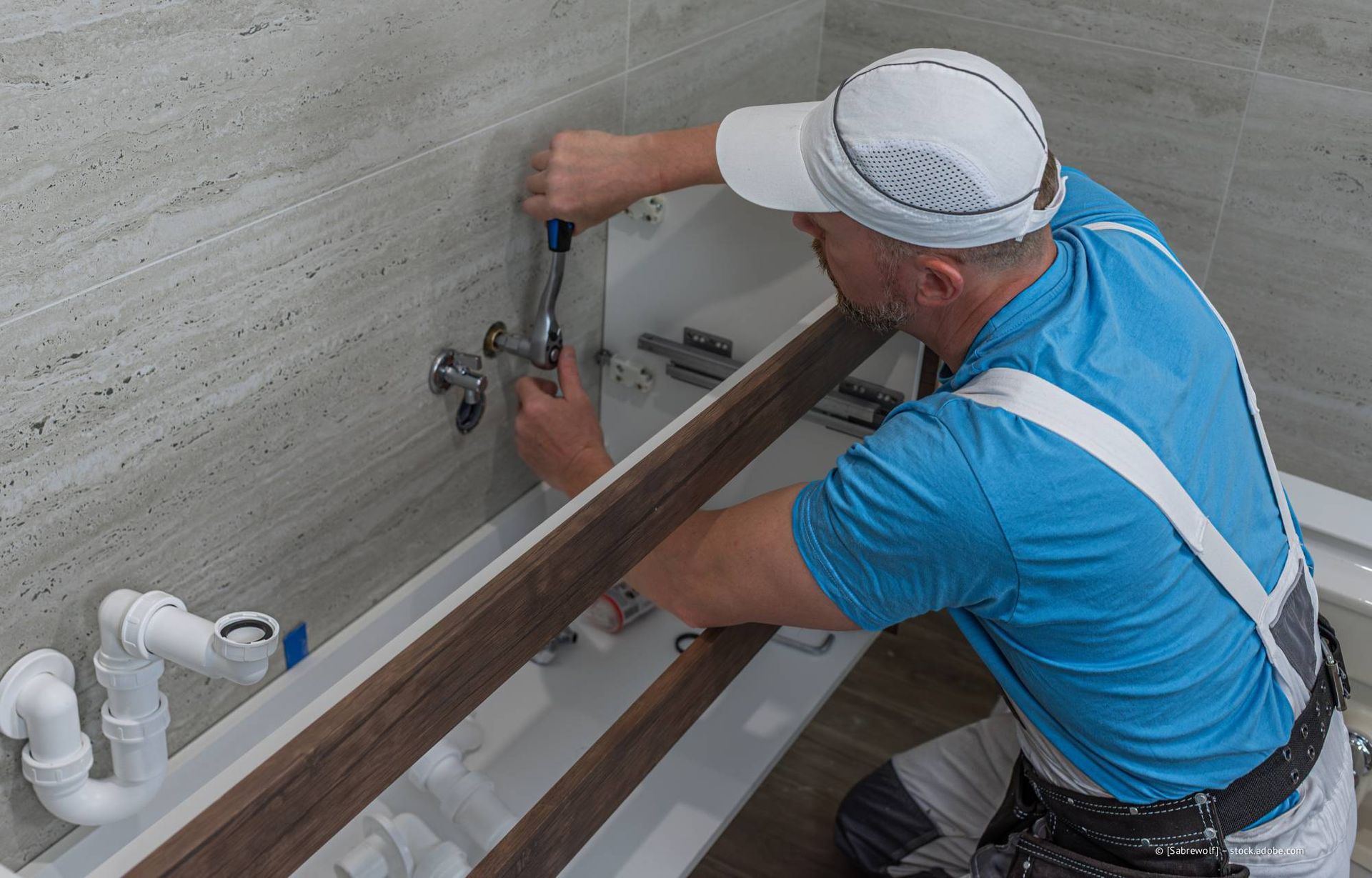 Ein Klempner arbeitet mit einem Schraubenschlüssel an den Rohren im Badezimmer. Er trägt eine Kappe und ein blaues Hemd.