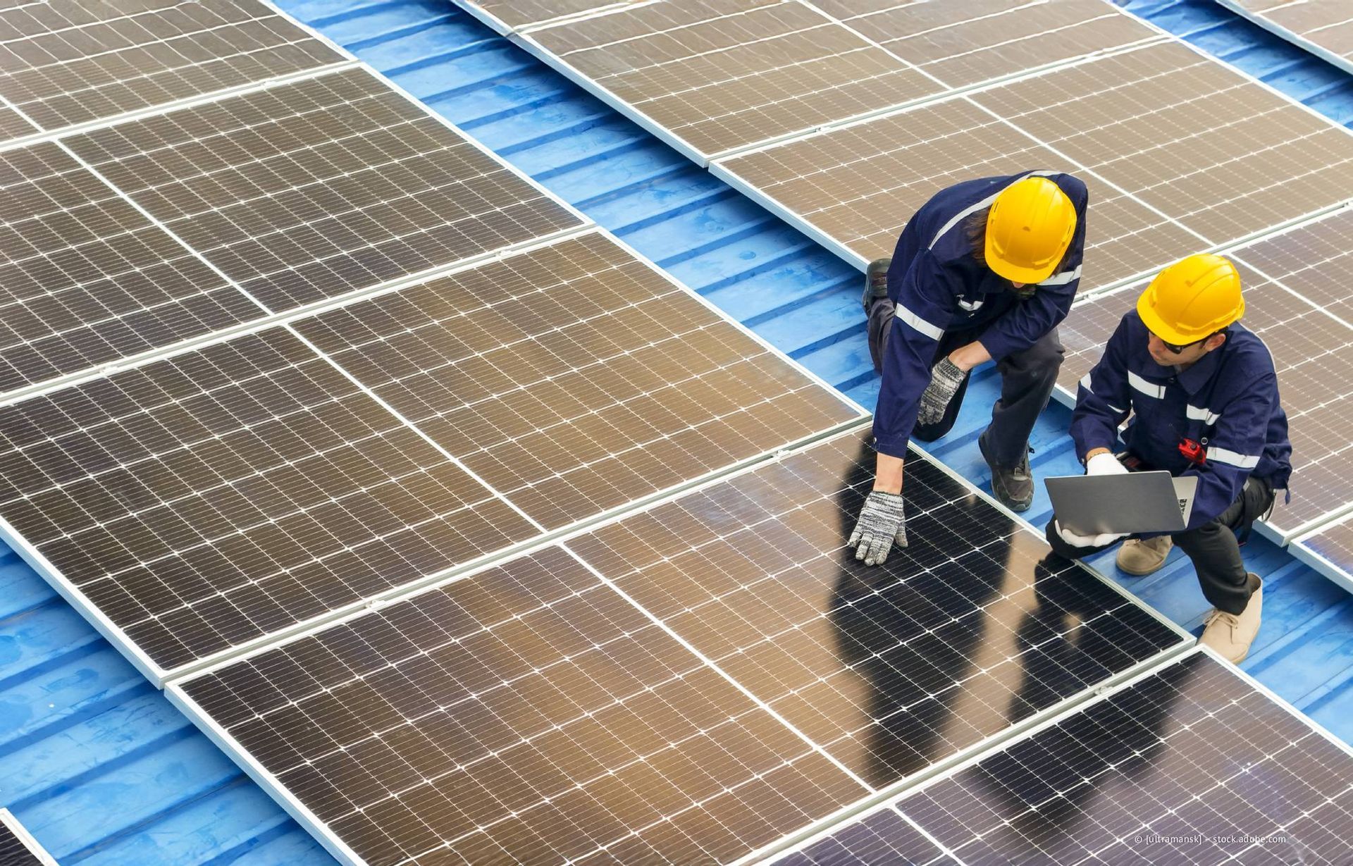 Zwei Arbeiter mit Schutzhelmen installieren Solarpaneele auf einem Dach.