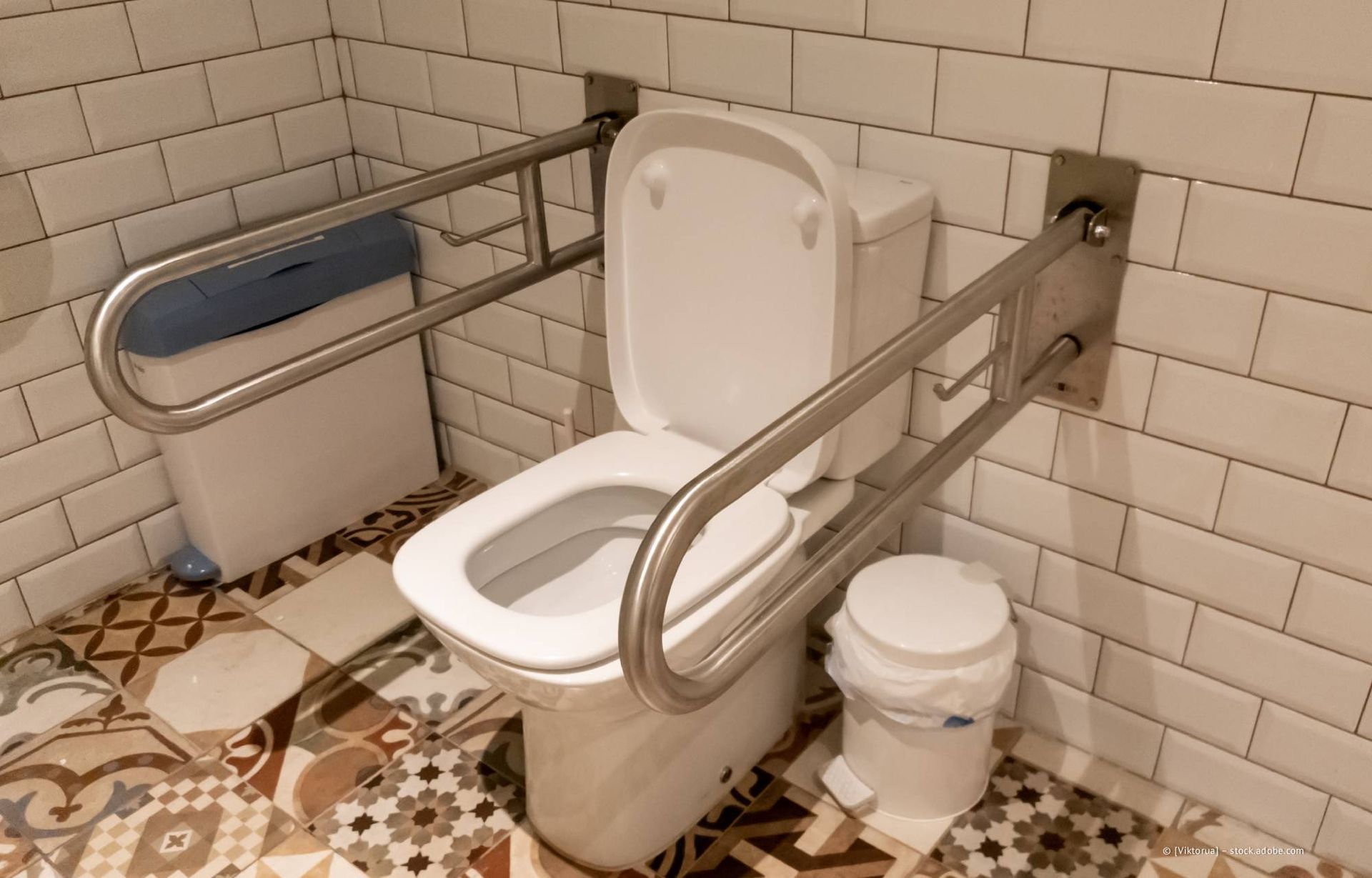 Toilette in einem gefliesten Badezimmer mit Haltegriffen an beiden Seiten und einem blauen Abfalleimer.