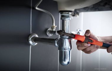Eine Person benutzt einen Schraubenschlüssel, um an einem Chromrohr unter einem Waschbecken in einem gefliesten Badezimmer zu arbeiten.