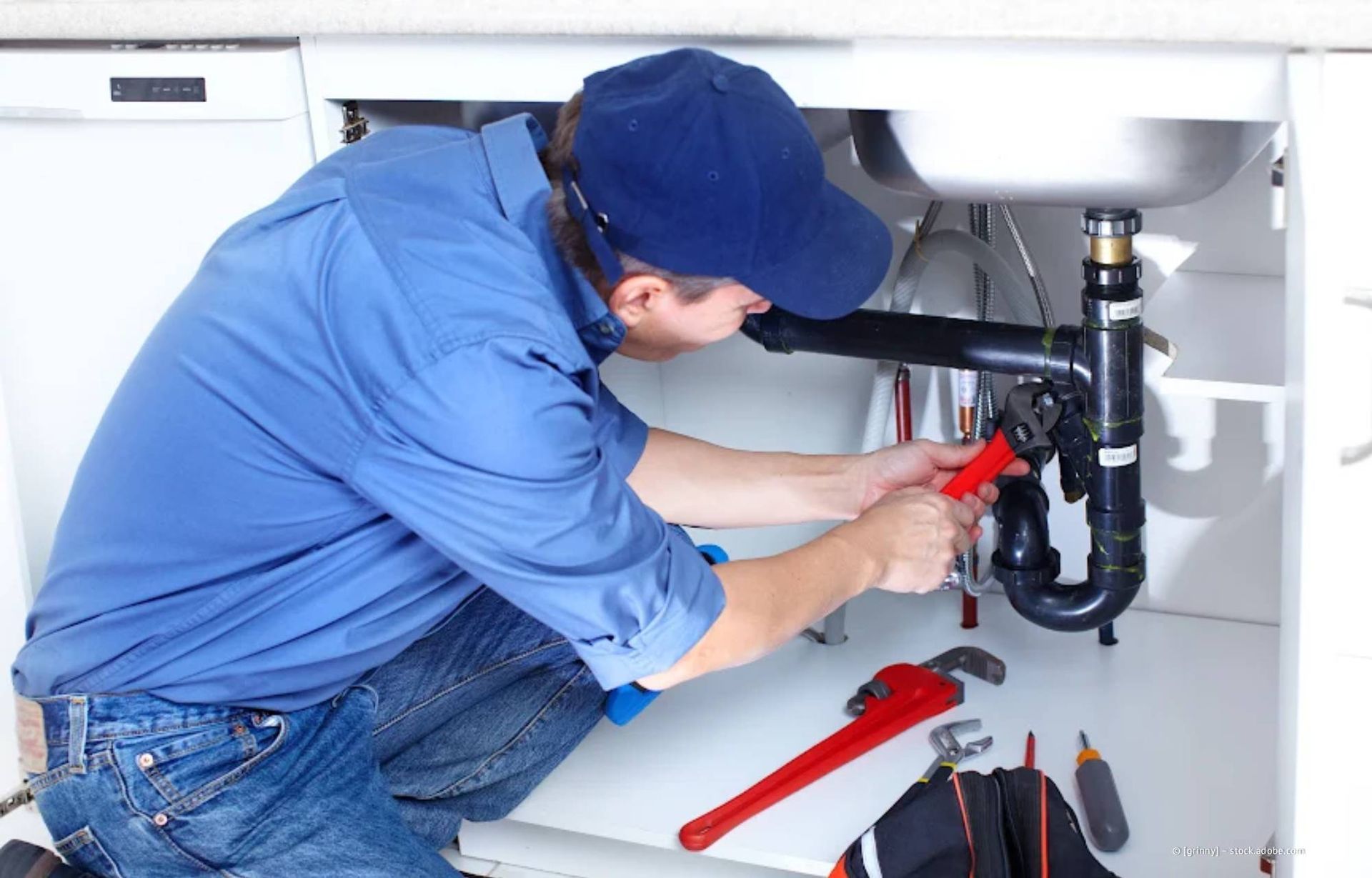 Ein Klempner in blauem Hemd und Kappe arbeitet mit einem Schraubenschlüssel unter der Küchenspüle.