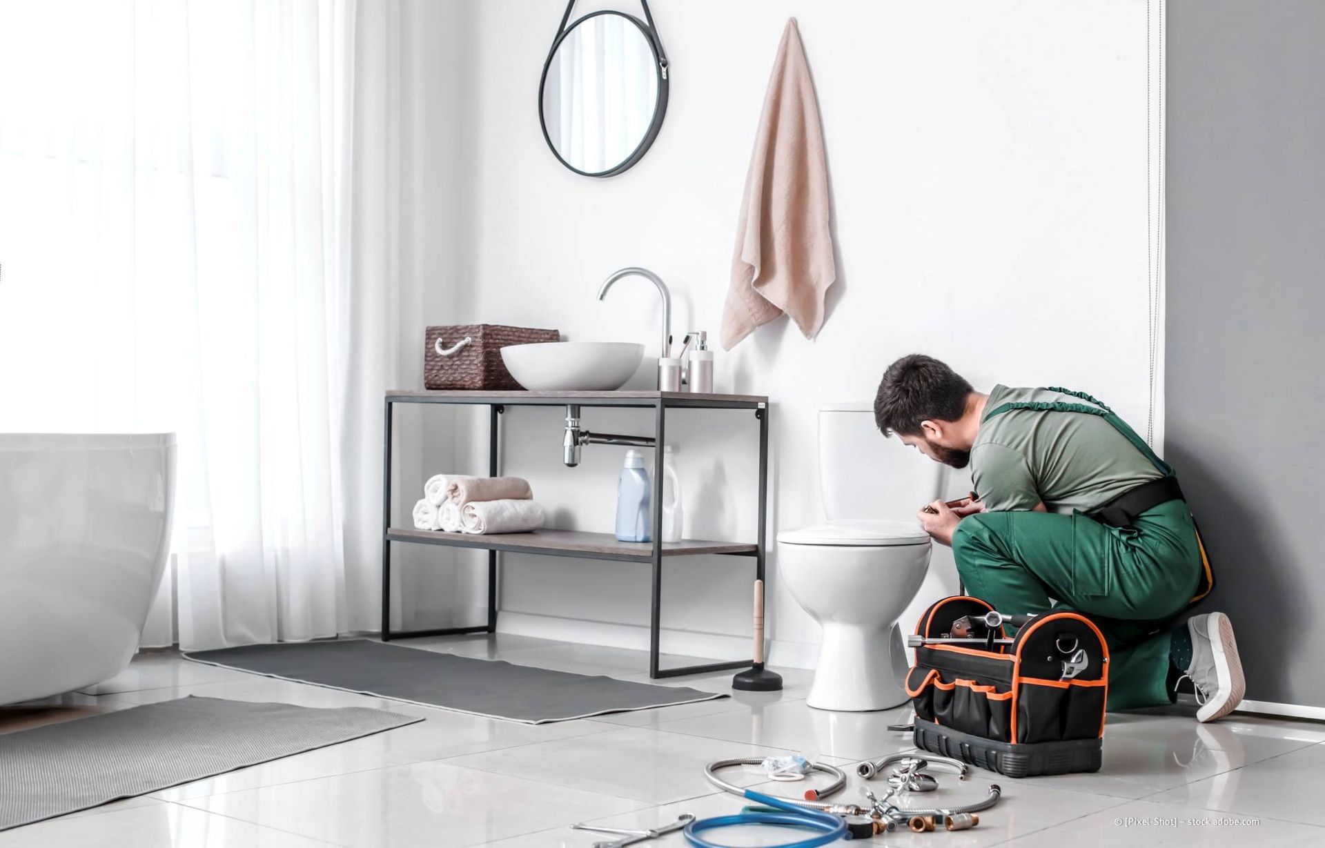 Ein Klempner arbeitet an einer Toilette in einem modernen Badezimmer. Werkzeugtasche auf dem Boden, weiße Wände und ein Waschbecken.