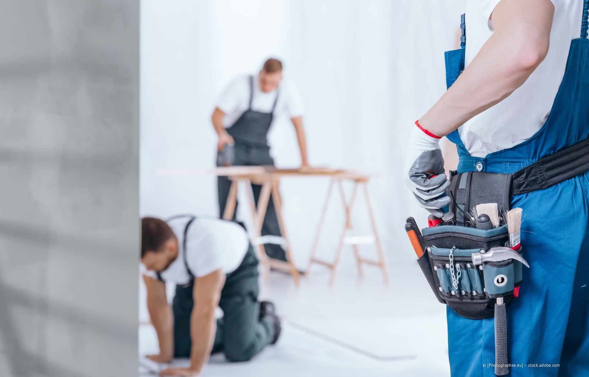 Bauarbeiter, einer mit Werkzeuggürtel, zwei weitere arbeiten an einem Holzprojekt in der Halle.