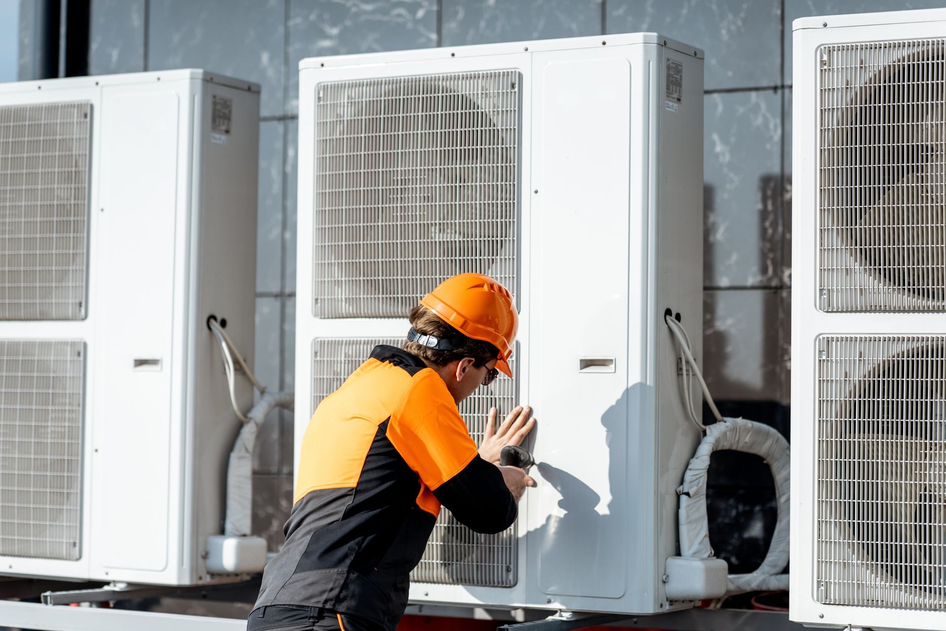 Un ouvrier portant un casque orange inspecte des unités de climatisation extérieures sur la façade d'un bâtiment.