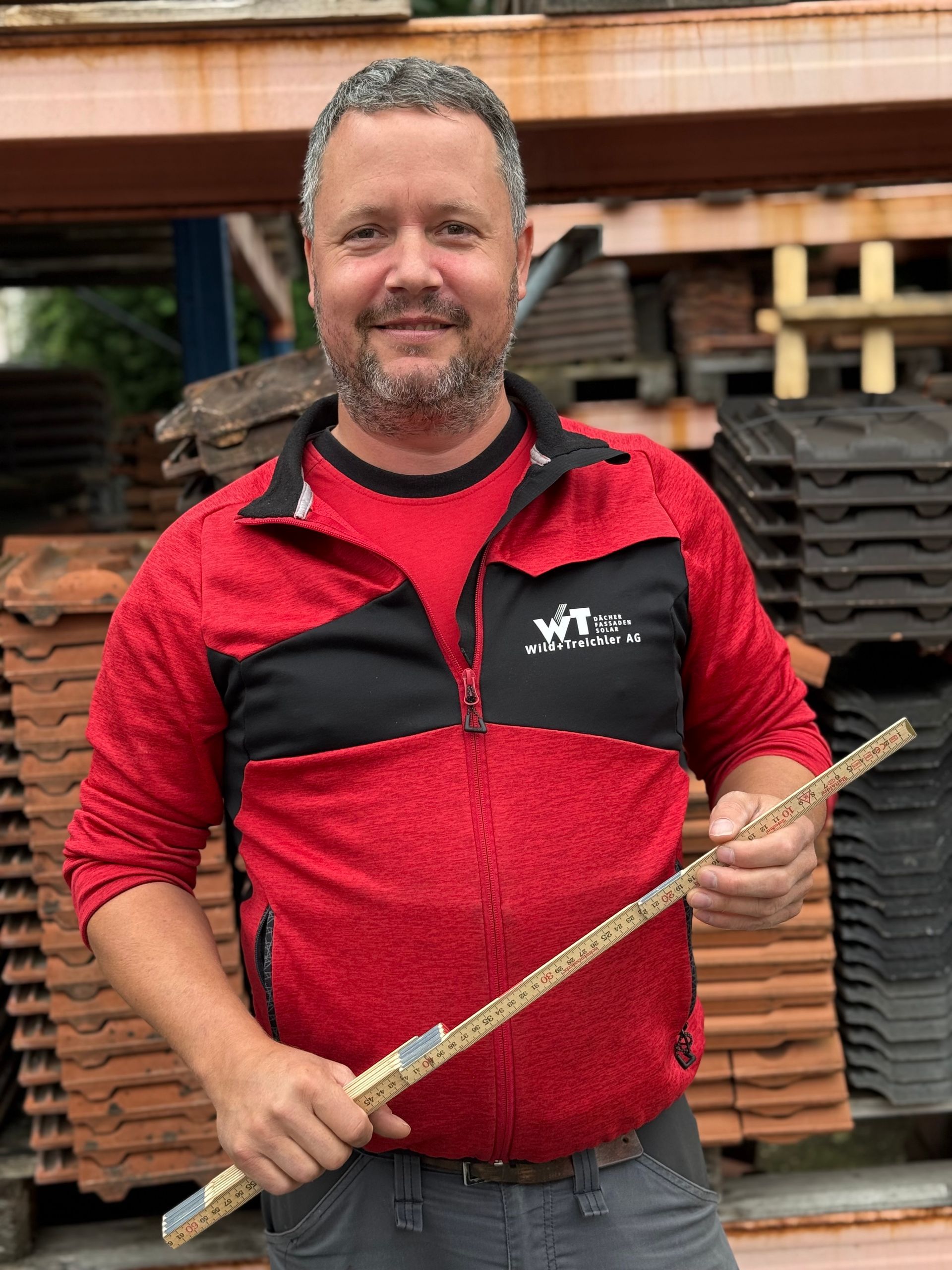 Mann in rotem Reißverschlussanzug hält einen Holzstab und lächelt. Steht draußen neben Stapeln von Dachziegeln.