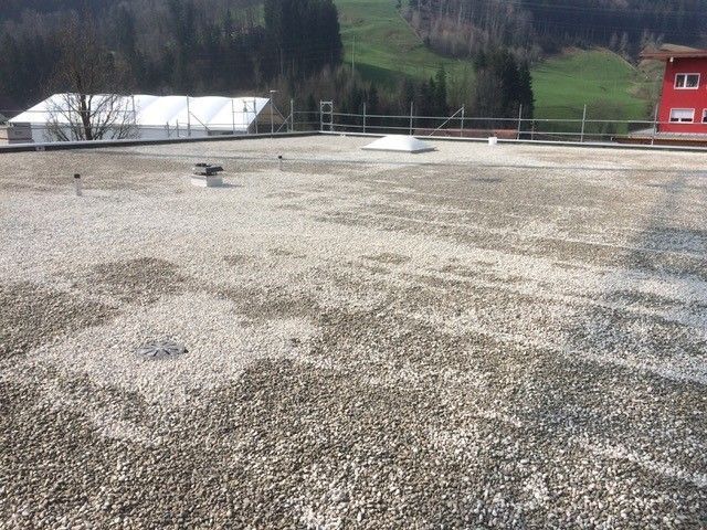 Flachdach mit Kies bedeckt, in der Ferne sind Hügel und ein rotes Gebäude zu sehen.
