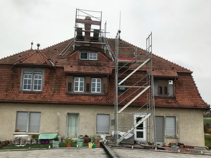 Arbeiter auf einem Gerüst reparieren das rote Ziegeldach eines hellen Gebäudes.