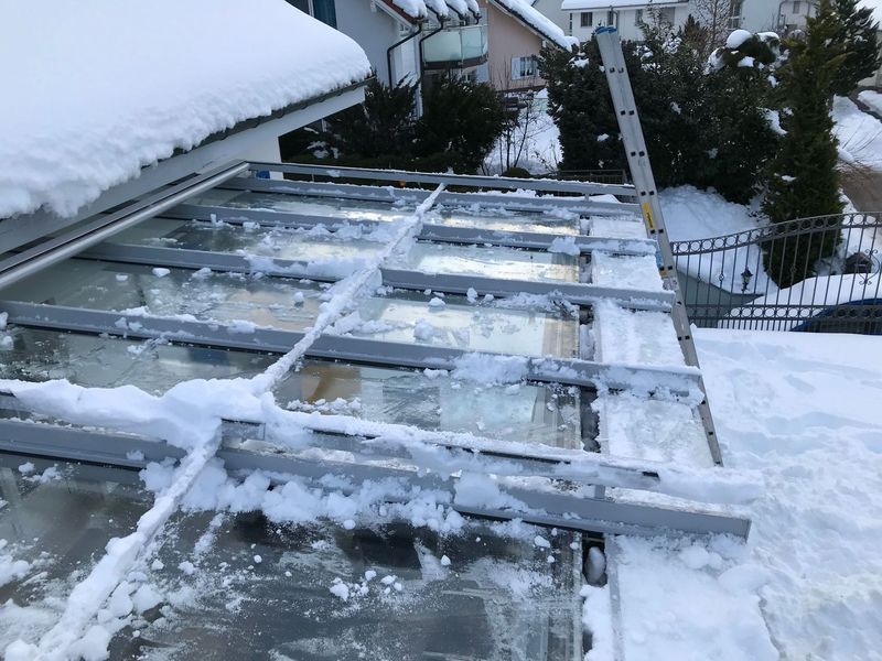 Schneebedecktes Glasdach mit Leiter, daneben ein schneebedecktes Haus.