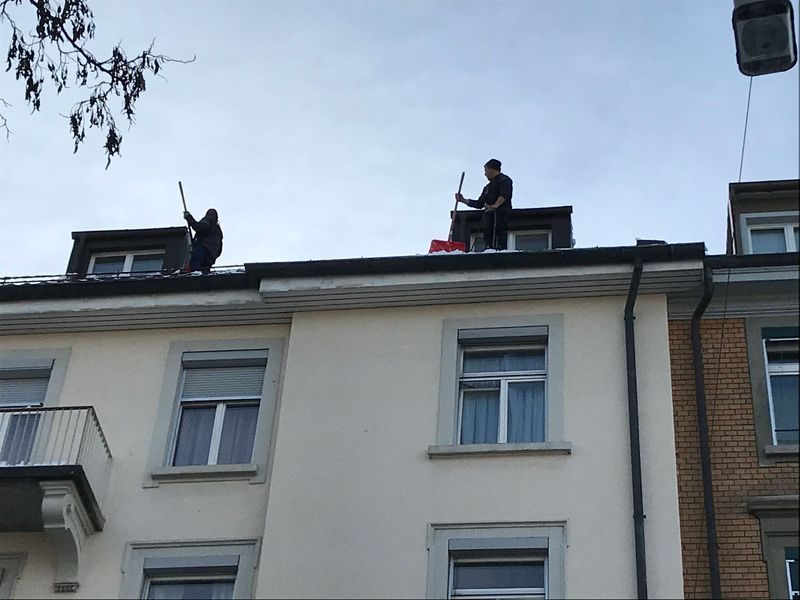 Zwei Personen räumen an einem bewölkten Tag mit Schaufeln Schnee von einem Gebäudedach.