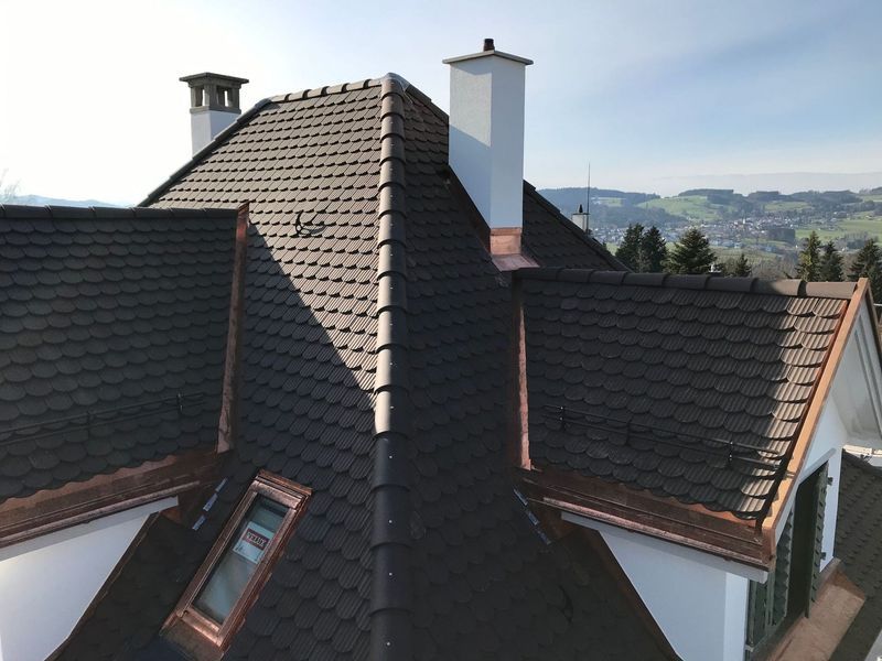 Dunkelbraunes Ziegeldach mit Kupferakzenten, Schornsteinen und einem Fenster auf einem weißen Haus mit malerischer Aussicht.