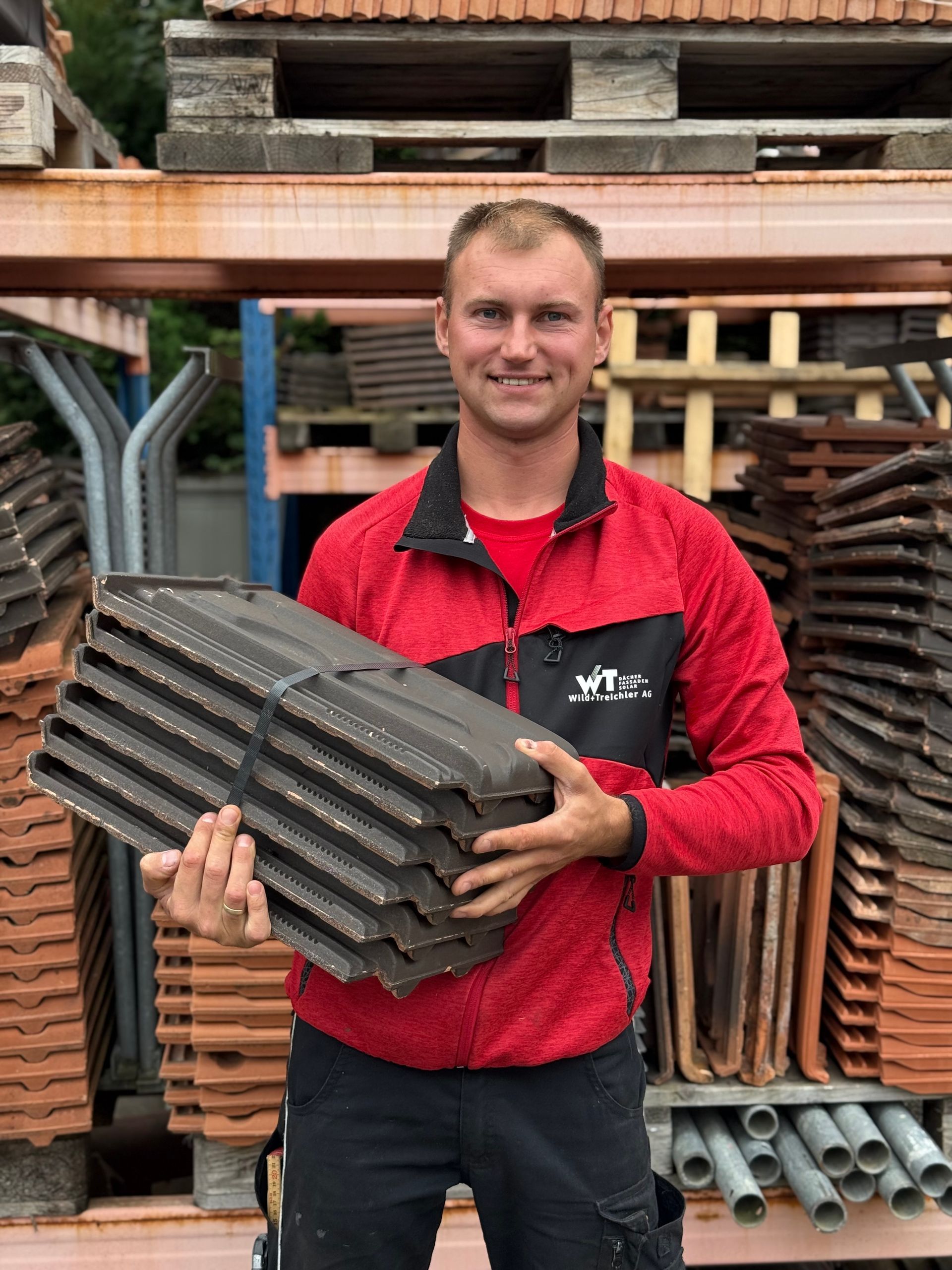 Mann in roter Jacke hält lächelnd Dachziegel auf einem Holzplatz.
