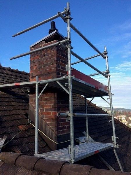 Ein Gerüst um einen gemauerten Schornstein auf einem Dach. Der Himmel ist blau.