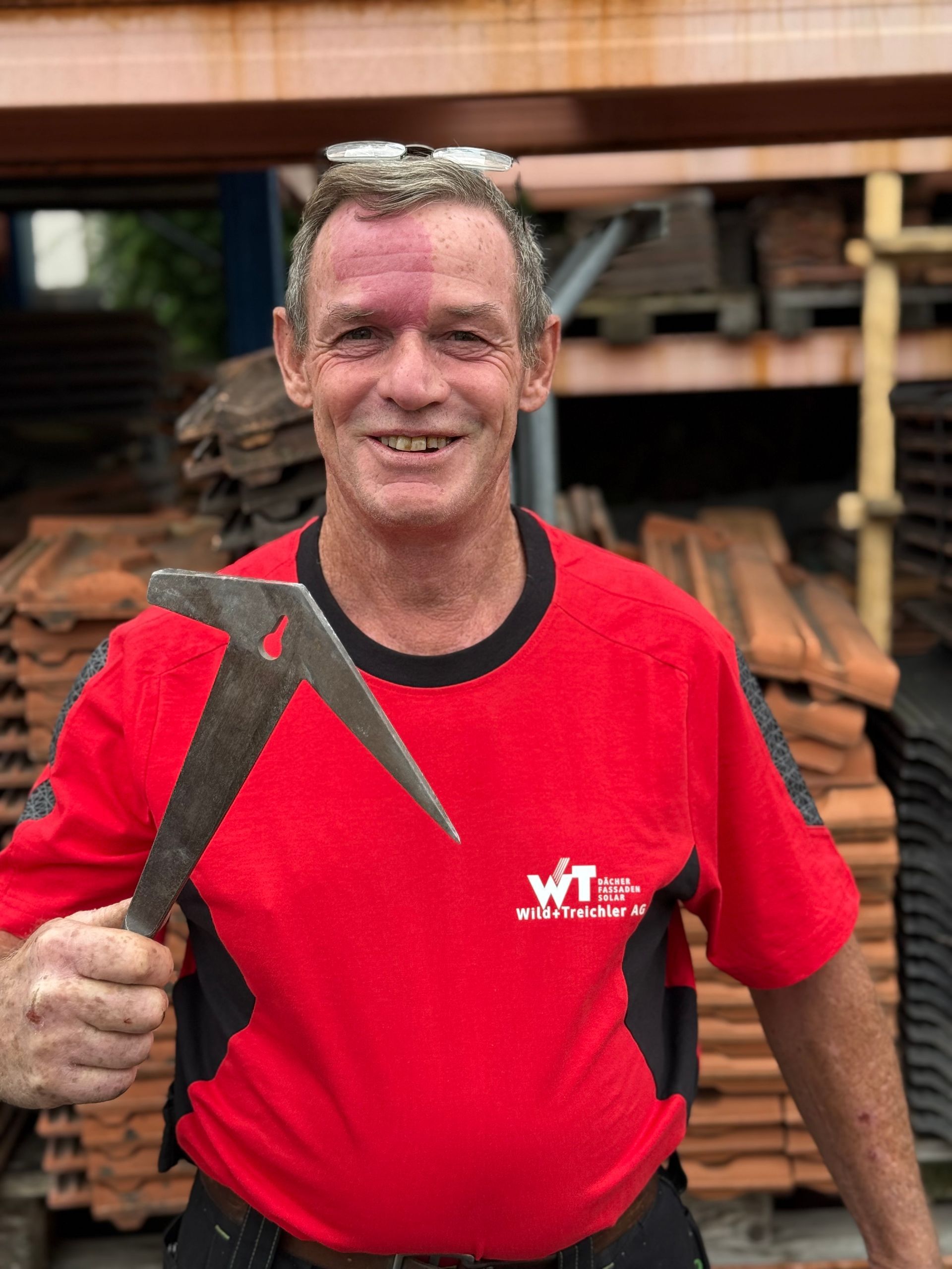 Mann im roten Hemd hält lächelnd ein Dachdeckerwerkzeug vor gestapelten Ziegeln.