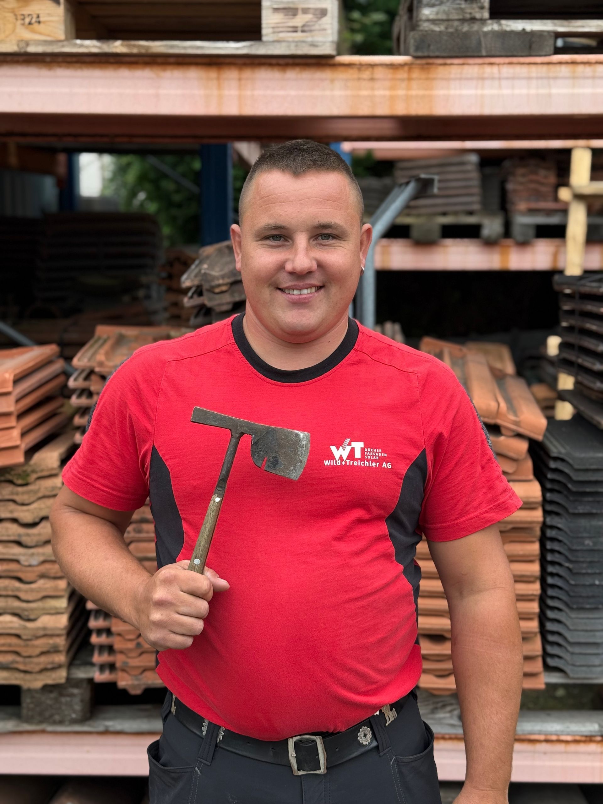 Mann im roten Hemd hält lächelnd einen Hammer in der Hand und steht vor Dachziegeln.
