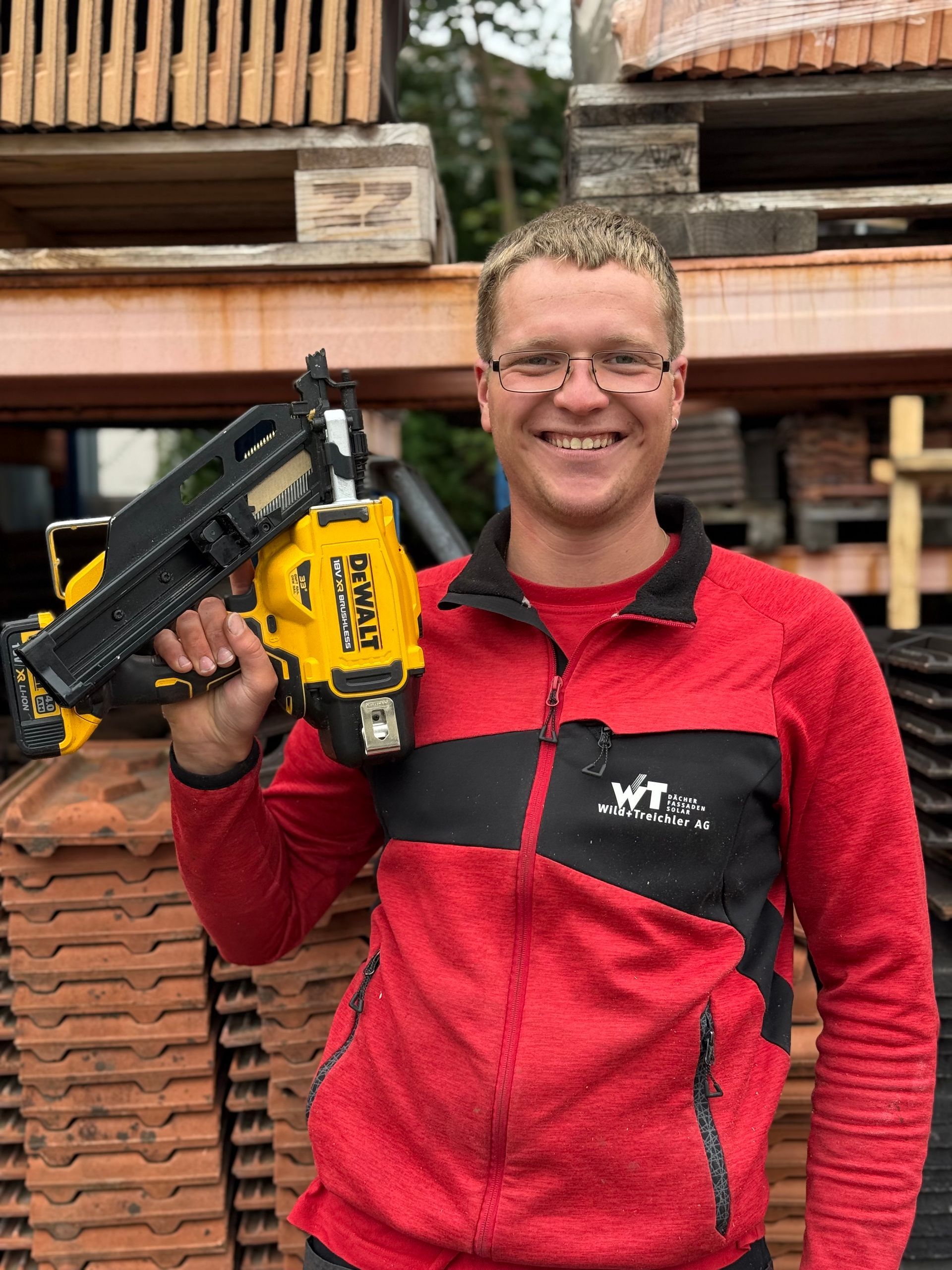 Mann in Rot hält lächelnd einen gelb-schwarzen Nagelschussapparat, umgeben von Baumaterialien.