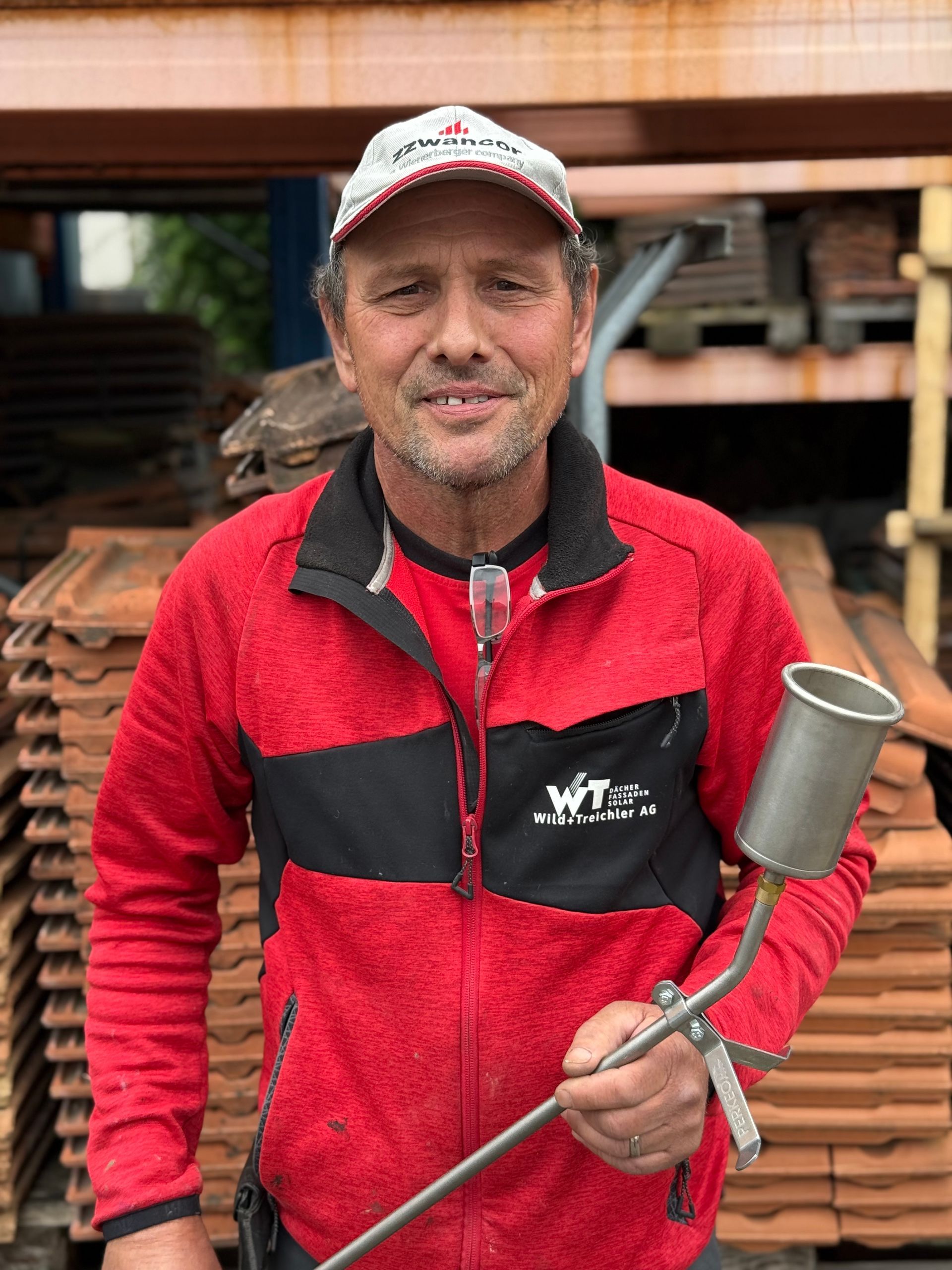 Mann in roter Jacke und Hut hält lächelnd ein Metallwerkzeug vor gestapelten Dachziegeln.