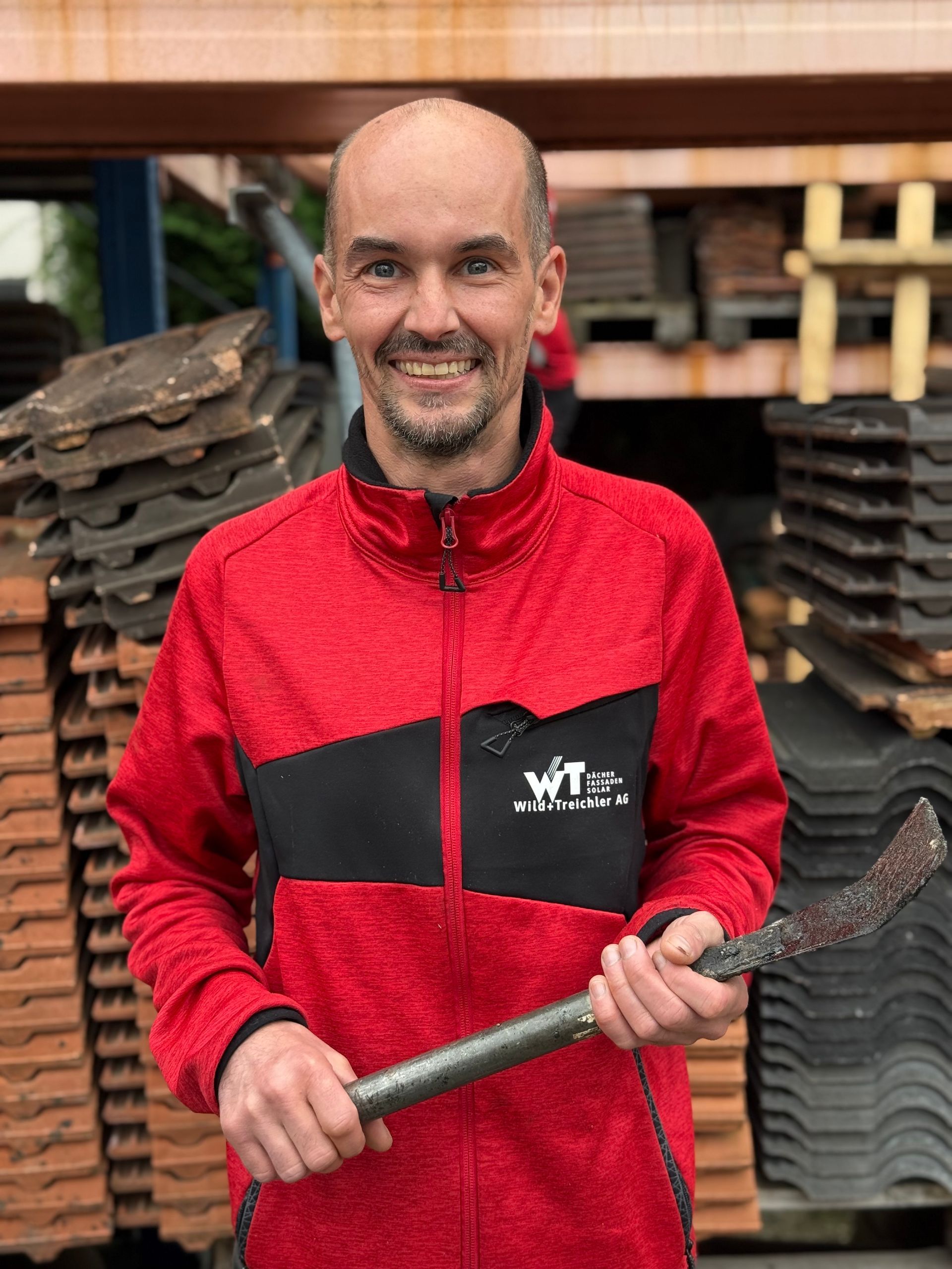 Mann in roter Jacke hält ein Dachdeckerwerkzeug und lächelt in einer Lagerhalle.