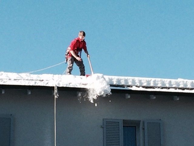 Person in roter Jacke schaufelt Schnee von einem Dach, gesichert durch einen Sicherheitsgurt, vor einem blauen Himmel.