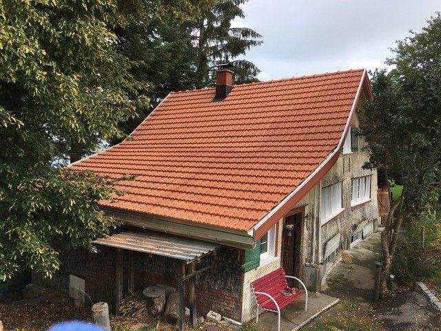 Kleines Häuschen mit rotem Ziegeldach, Schornstein und einer roten Bank davor, umgeben von Bäumen.