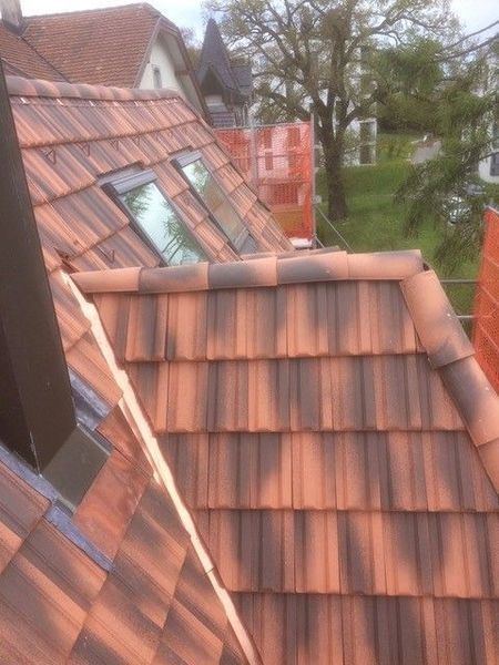 Nahaufnahme eines braunen Ziegeldachs mit zwei Oberlichtern. Im Hintergrund sind Bauarbeiten im Gange.
