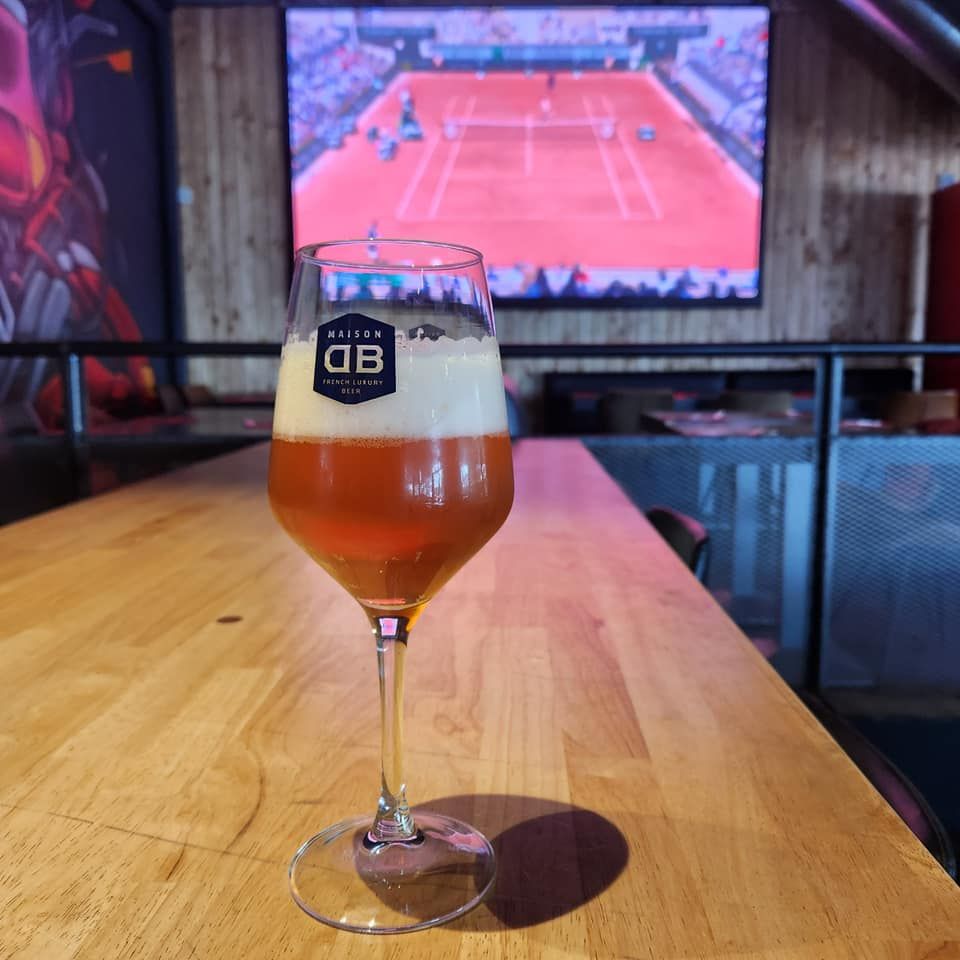 Un verre de bière sur une table en bois, avec un match de tennis diffusé sur un écran en arrière-plan.
